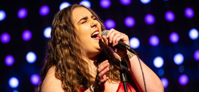 A student with long brown curly hair sings into a microphone.