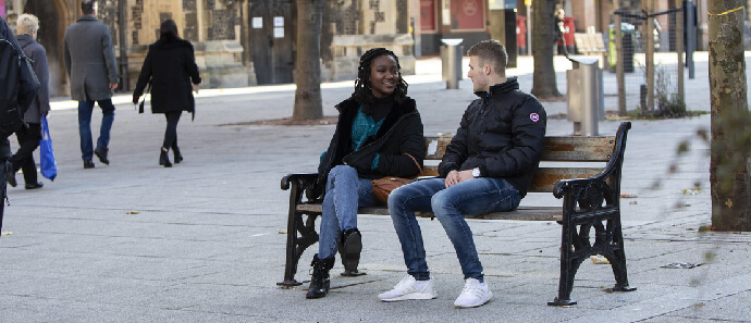 Two students, one male and one female, sit on a bench talking in Reading town centre.